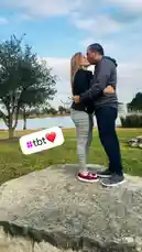 A couple shares a tender kiss while standing together on a large rock in a scenic park setting. The woman wears a black top and gray leggings, while her partner is dressed in a gray hoodie and dark pants. They embrace affectionately under a partly cloudy sky, with a calm lake and trees visible in the background. A graphic overlay reading '#tbt ❤️' suggests this is a throwback photo, capturing a cherished memory. by @kastielagarcia with kastielagarcia by Contains: Amor Bonito - Kany García & Juan Luis Guerra
