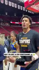 Two players from the NBA Summer League champions team celebrate their victory on the court in Las Vegas. They are seen wearing black 'CHAMPIONS' t-shirts and holding their championship trophies, including a large circular trophy and rings in black boxes. The atmosphere is festive, with fans in the stands and banners confirming the event as the 'NBA SUMMER LEAGUE CHAMPIONS VICTORY IN LAS VEGAS'. by @nba with Original Sound by nba