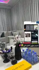 A stylish and modern living room setup featuring a white sofa, elegant curtains, and a coffee table adorned with a black vase of white flowers, a tray of water bottles, a yellow box labeled 'HAVANA', and decorative items. The ambiance is enhanced by soft lighting and a contemporary design aesthetic. by @batoolmakeup1 with original sound