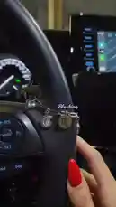 A close-up shot inside a car focuses on a stylish charm bracelet attached to the steering wheel, featuring zodiac signs and a heart. A hand with red nail polish gently holds the wheel. Overlaid text reads 'Blushing'. In the background, a man speaks in Arabic, saying, 'Allah loves you, and your love doesn't decrease with time; it increases in your absence.' This intimate scene combines personal style with a heartfelt message. by @bx301a with 🎻🎻📻 by 7bk