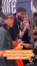In this behind-the-scenes look, NBA player Mikal Bridges is seen clocking in for his shift as a cashier at the official NBA store. Dressed casually in a black shirt and backwards cap, he interacts with a fellow employee who is scanning merchandise. A fan captures the moment on her phone, smiling as Bridges handles a bright orange t-shirt. The scene provides a rare glimpse into the player's off-court life and community engagement. by @yahoosports with Original Sound by yahoosports