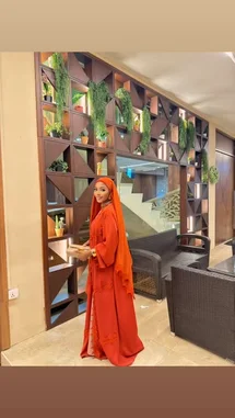 In this stylish moment, Everything Naddy showcases a stunning red hijab and matching traditional outfit in a chic, modern interior. The setting features a unique geometric wall with lush greenery and contemporary wicker furniture, creating a perfect backdrop for her look. by @everythingnaddy with she go by💃 by Contains: she goes by. (KayArchon Remix) - KayArchonn & where.t.at