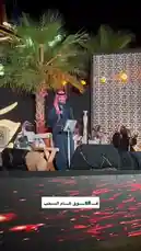 Experience the vibrant atmosphere of a live traditional music performance at night during an 'إيفنت جَلسة' (Jalsah event). A central performer sings into a microphone while musicians play instruments like drums and wind instruments behind him. The stage is illuminated by warm lights against a backdrop of palm trees and a decorative wall, with dynamic red lighting effects projected onto the floor. by @jalsahevent with jalsahevent by Original Sound