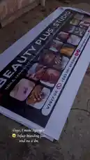 A close-up view of a large advertising banner for Beauty Plus Studio, laid out on a concrete floor. The banner prominently displays the studio's name, contact information, and images showcasing services like nails, lashes, tattoos, and piercings. by @naa.ayele911 with Original Sound by naa.ayele911