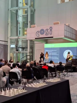 A live event hosted by Chess.com features a panel discussion on stage with multiple speakers addressing an audience. In the foreground, several chessboards are set up on tables, suggesting a simultaneous exhibition or tournament. The modern venue includes a prominent 'cosi' sign, and a large screen displays presentation slides behind the speakers. by @chesscom with original sound
