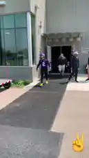 A player wearing a purple Minnesota Vikings jersey with the number 2 walks out of a building and onto a practice field. He makes a peace sign gesture towards the camera before continuing his walk across the grass. The setting appears to be a sunny day at a team facility. by @vikings with original sound