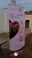 A banner for the 'Peace Message Volunteer Team' is displayed outdoors, announcing a new initiative with the Arabic text 'We are launching...'. The banner features a heart-shaped photo of volunteers in action, a QR code for donations, and logos from partner organizations, including the Red Crescent. by @peace.message with original sound