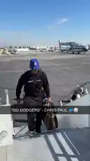 A man, identified as an LA Clippers player, confidently walks down the stairs of a private jet on a sunny day. He is wearing a black 'KEEP SMILIN' sweatshirt, a blue 'LA' baseball cap, and carrying a designer handbag, showcasing a casual yet fashionable airport look. by @clippers with Original Sound by clippers