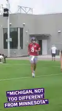 A player wearing a red jersey with the number 8 and a purple helmet walks onto a grassy field during what appears to be a practice session. In the background, other players can be seen, and a modern building stands behind the field. A text overlay reads 'MICHIGAN, NOT TOO DIFFERENT FROM MINNESOTA,' suggesting a comparison between the two states. by @vikings with Original Sound by vikings