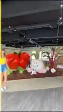 In this playful scene, Martine Beerman interacts with a whimsical indoor garden display featuring giant, cartoonish vegetables like tomatoes, onions, and beets. She strikes a pose with her arms outstretched, creating the illusion of holding the oversized produce. The background includes a dirt patch with small plush characters, adding to the imaginative atmosphere. by @martine with Island In The Sun by Weezer