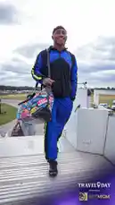 A smiling Baltimore Ravens player walks down the ramp from an airplane, carrying a large bag covered in travel stickers. He's dressed in a blue and black Under Armour tracksuit, showcasing team pride during a 'Travel Day' event presented by BetMGM. This moment captures the anticipation and excitement before a big game. by @bltravens with original sound