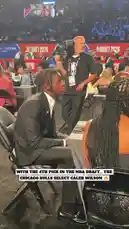 A young man in a suit is shown sitting anxiously at a table during the NBA Draft 2026. As his name is announced, he stands up, embraces people beside him—including a woman in a black dress—and then walks onto a stage to shake hands with an official. The event takes place in a large arena with 'CHICAGO BULLS' branding visible on the stage. by @nba with Original Sound by nba