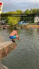 A shirtless man in blue swim trunks prepares and then leaps off a large rock into a lake below, creating a massive splash. He resurfaces moments later as a bright orange object floats nearby. The scene takes place at a lakeside park with people, tents, and a white house visible in the background under a cloudy sky. by @digiflips with Original Sound by digiflips