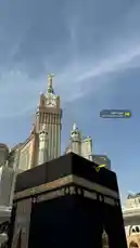A serene, upward-looking view captures the majestic Kaaba draped in its black kiswa, set against the backdrop of the towering Abraj Al-Bait complex and its iconic clock tower. The clear blue sky enhances the grandeur of these holy sites, creating a powerful and peaceful visual. by @batoolmakeup1 with سورة الكهف by Sheikh Fahad AlKandary