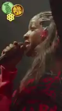 A vibrant performance by Sabrina Carpenter at the Big Chill Festival 2024. She sings passionately into a microphone on stage, dressed in a red and black patterned top with pink sleeves, a studded belt, and long dark hair adorned with clips. The audience is visible in the foreground, recording the show on their phones under atmospheric stage lighting. by @daisychainfield with Original Sound by daisychainfield