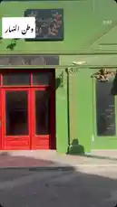 A stylish woman wearing a beige coat and white headscarf confidently walks past a striking green building with red doors. The scene captures urban elegance and modern fashion in motion. by @artal.street with artal.street by Original Sound