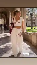 A young woman walks through a sunlit university courtyard, showcasing a trendy 'SHARK' themed outfit. She wears a white crop top and matching sweatpants, carrying a red tote bag and a book, all while smiling. This scene, associated with the TikTok user 'Mark The Shark', captures a moment of campus life and fashion. by @mts550 with original sound