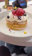 A close-up, step-by-step look at creating a beautiful stack of pancakes topped with whipped cream, fresh strawberries, blueberries, and edible glitter. The camera focuses on the final presentation on a white plate, set against a stylish table setting with a phone displaying a music playlist, suggesting a perfect morning treat. by @miji-bello with Original Sound by miji-bello