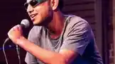 This high-energy performance features K-DEV, a charismatic singer wearing a gray t-shirt and distinctive green-framed sunglasses. He passionately sings into a microphone, his expressive face and dynamic posture conveying deep emotion and connection with the music. The intimate setting, likely a small concert or open mic night, highlights his raw talent and stage presence. by @k_dev170 with Original Sound by k_dev170