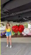 In a vibrant indoor setting filled with oversized, whimsical vegetable props like a giant tomato and a smiling radish, Martine Beerman strikes playful poses. She wears a bright yellow tank top and denim shorts, dancing and gesturing enthusiastically in front of the colorful backdrop. by @martine with Yes by LMFAO