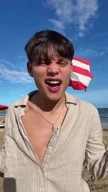 In this bright, sunny beach scene, S E B A S 🧸 stands against a backdrop of blue sky and ocean waves. Wearing a light-colored button-up shirt and a necklace, he makes direct eye contact with the camera, smiling warmly and gesturing expressively with his hands. A red and white striped flag flaps in the wind behind him, adding a dynamic element to the relaxed coastal atmosphere. by @twinksebass with Original Sound by Contains: Nicole Kidman - ADÉLA