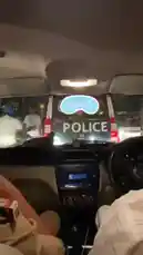 This first-person view shows the interior of a police car during a night patrol. An officer in uniform sits in the driver's seat, holding a smartphone, while another person occupies the passenger seat. Through the windshield, the illuminated 'POLICE' markings on the front of the vehicle are visible against the dark background. by @im.navnit with Police by Cheema Y, Gur Sidhu, Jasmeen Akhtar
