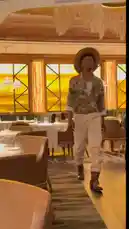 A stylish individual, identified as Delly, confidently strides through a modern restaurant. They are wearing a distinctive wide-brimmed hat, a patterned jacket over a white shirt, and light-colored pants. The warm, ambient lighting highlights their unique fashion sense as they walk down the aisle. by @delwinbazilme with World Cup by iShowSpeed