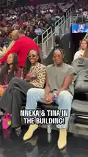 Two women, identified by on-screen text as Nneka and Tina, are seen sitting together in the stands at a crowded indoor sports arena, likely during a WNBA game. They are smiling and looking towards the camera. One woman waves her hand. The background shows other spectators and a large screen displaying a logo. by @wnba with Original Sound by wnba