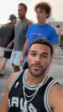 A young man, identified as Jeremiah Brown, films himself and friends walking down stairs in a black San Antonio Spurs jersey. The group then moves through a sunny, palm-lined downtown area, passing a prominent 'Downtown! Oceanside' sign. The audio captures the excitement of a Spurs fan, creating a casual vlog-style moment. by @findjeremiah with Original Sound by findjeremiah