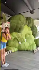 In this viral social media moment, influencer Martine Beerman performs a classic optical illusion. Wearing a yellow top, denim shorts, and a cute bunny hat, she stands next to a massive, green broccoli sculpture. By extending her arm perfectly parallel to the ground, she creates the illusion that she is touching the giant vegetable, which is actually much further away. The scene takes place indoors, possibly in a modern office or creative space, adding to the surreal and playful atmosphere. by @martine with Eat Your Veggies by Doggyland