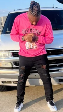 In this eye-catching scene, rapper PLAYBOII Q stands confidently in front of a silver SUV, showcasing his bold fashion sense. He wears a vibrant pink long-sleeve graphic tee with 'BIZARRE' printed across the chest, paired with heavily distressed black jeans and crisp white sneakers. His signature braided hairstyle adds to the distinct aesthetic. The bright daylight and urban backdrop highlight his unique style, making it a standout moment for fans and fashion enthusiasts alike. by @quezz.706 with original sound