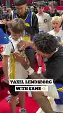 A professional basketball player from the Golden State Warriors kneels down on the court to sign autographs for young fans wearing team jerseys. The scene captures a heartwarming moment of athlete-fan interaction, with other children and adults watching nearby in a crowded arena setting. by @nba with Original Sound by nba