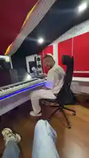 A first-person perspective tour of a modern music production studio. A man, identified as Delly, is seen working at a large keyboard setup with LED lighting. The camera pans around the room, showcasing professional audio equipment, including speakers and a microphone, all within a space featuring red acoustic panels and a stylish black-and-white ceiling design. by @delwinbazilme with Original Sound by delwinbazilme