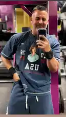 In this selfie-style clip, fitness personality Mark The Shark takes a mirror photo inside a gym. He's wearing a blue t-shirt with a graphic celebrating 'USA' and a portrait of Sabrina Carpenter, paired with matching sweatpants. The setting features purple gym equipment and bright lighting, capturing a moment where personal fitness meets pop culture. The image suggests themes of health, self-expression, and contemporary celebrity influence. by @mts550 with original sound