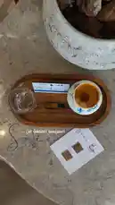 A beautifully arranged espresso coffee moment is showcased on a wooden tray atop a marble surface. The scene features a small cup of espresso, a glass of water, a sugar packet, and a decorative card with Arabic text reading 'الجمعة من ... اسبريسو' ('Espresso from Friday...'). A large ceramic pot sits nearby, adding to the cozy ambiance. This elegant setup, attributed to كريّم 📿, evokes themes of café culture, relaxation, and aesthetic presentation. by @karim.alsaleh with original sound