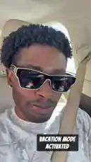 A close-up, selfie-style shot captures Delly inside a car, showcasing his curly hairstyle and trendy white-framed sunglasses. He is wearing a light-colored, patterned t-shirt and has a seatbelt fastened. The interior of the vehicle is visible around him. Throughout the clip, he subtly moves his head and shifts his gaze, maintaining a calm expression. The audio features a modern hip-hop track with a male vocalist rapping over a steady beat. by @delwinbazilme with Backwards by Quavo, T.I.