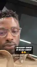 A close-up selfie from Delly at Concourse A in Atlanta, GA. He's wearing clear-framed glasses and a tan shirt, looking directly into the camera with a neutral expression. The background shows the typical airport terminal lighting and architecture. by @delwinbazilme with OMG by Usher feat. will.i.am