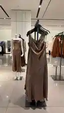 A luxurious gold-toned maxi dress with a draped neckline and spaghetti straps is showcased on a black hanger in a well-lit retail store. In the background, a mannequin wears a similar shimmering brown dress, highlighting a cohesive collection of elegant evening wear. The modern interior features polished floors and contemporary fixtures, creating a sophisticated shopping ambiance. by @ru.ab71 with original sound