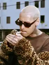 In this close-up shot, JeZ Dior is seen wearing dark sunglasses and a brown shirt, wrapped in a striking leopard print coat. He holds a cigarette near his mouth, showcasing visible tattoos on his neck and hands. The setting appears to be outdoors during the day, with a building in the background. His shaved head features a small tattoo above his right eyebrow, adding to his distinctive appearance. This stylish moment captures a unique blend of fashion, attitude, and personal expression. by @jezdior with original sound