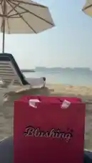 A vibrant pink shopping bag from 'Blushing The charm boutique' takes center stage on a pristine sandy beach. In the background, a lounge chair and umbrella offer a relaxing seaside view, while upbeat Latin music plays, setting a festive summer mood perfect for a day out. by @bx301a with 🕺🏻🕺🏻🕺🏻🕺🏻🕺🏻🕺🏻🕺🏻🕺🏻🕺🏻 by As