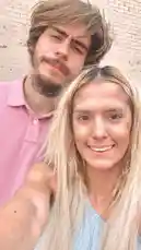 In this charming selfie, Kaylee shares a sweet moment with her boyfriend. They are standing close together, smiling warmly at the camera against a white brick wall. Kaylee, with long blonde hair, is holding the camera to capture the shot, while her boyfriend, wearing a pink polo shirt, looks on with a gentle expression. The overall mood is happy and affectionate. by @csnwkf1 with Original Sound by Contains: Can't Break Up Now - Old Dominion & Megan Moroney