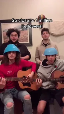 In a joyful celebration of nearing one million followers, the AMH Band performs a playful musical challenge. Two members strum acoustic guitars while wearing bright blue plastic helmets, adding a comedic twist to their performance. The other two members provide energetic vocal support and rhythmic clapping, creating a lively and entertaining atmosphere perfect for social media engagement. by @amhbande with amhbande by Original Sound