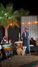 A captivating live performance of traditional Arabic music unfolds on an outdoor stage at night. A male singer, dressed in a dark thobe and red-and-white ghutra, stands at a lectern singing into a microphone. Behind him, musicians in white thobes play traditional instruments like the oud and percussion. The stage is beautifully lit with warm lights against a decorative geometric backdrop, flanked by palm trees, creating an authentic and festive atmosphere. by @jalsahevent with jalsahevent by Original Sound