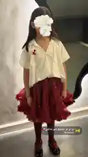 This video showcases a stylish school uniform look created by Ratib and More. A young girl models a chic outfit featuring a classic white button-up shirt paired with a modern, layered red tulle skirt adorned with subtle sparkles. The ensemble is completed with matching red knee-high socks and black Mary Jane shoes. The look is accessorized with a large, elegant white flower hair clip and a small red pin on her shirt, blending traditional school attire with contemporary fashion elements. by @om.louraa with لو لو لو اشيلج على كتافي🇰🇼 by Mykuwait
