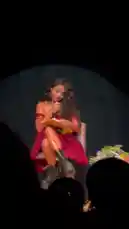 A woman with long dark hair sits on a stool under a spotlight on a stage, reading from a notebook into a microphone. She is wearing a red dress and black boots. The audience is visible in silhouette in the foreground. by @squashha with squashha by Original Sound