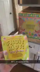 A hand holds a brightly colored box of Fruit Riot sour grapes candy, showcasing its 'real fruit, unreal flavor' tagline. In the background, a larger, festive Fruit Riot storage container sits on a kitchen counter. A text overlay reads 'this is all i survive on btw,' suggesting a humorous take on snack preferences. by @jezdior with original sound
