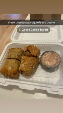 In this quick foodie snapshot, we see a close-up of crispy, golden-brown cheesesteak egg rolls served with a side of dipping sauce in a takeout container. The text overlay reads 'these cheesesteak eggrolls are bussin' and tags the location as 'Ocean Casino Resort.' This enticing treat highlights a popular fusion snack enjoyed at the resort. by @dimpled.diva02 with original sound
