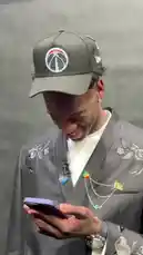 A stylishly dressed NBA player, wearing a Washington Wizards cap and a floral-embroidered suit jacket, is seen looking at his phone and smiling during a candid moment. He receives a message about an opportunity and acknowledges it, while being aware of the surrounding cameras. This appears to be a behind-the-scenes glimpse into the life of a professional athlete. by @nba with Original Sound by nba
