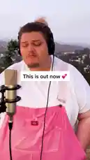 In a stunning outdoor performance overlooking a sprawling cityscape, musician TALK passionately sings their new release, 'This Is Out Now'. Wearing a white t-shirt and vibrant pink overalls, they deliver emotional lyrics like 'Where did I go wrong? I lost a friend somewhere along in the bitterness' into a professional microphone. The video features text overlays promoting the song's availability and thanking TikTok for its support, culminating in a call to action for viewers to find the link in the bio. by @iamtalk with iamtalk by Contains: How To Save A Life - TALK