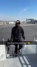 A man, identified as an LA Clippers player, confidently walks down the stairs of an airplane on a sunny day. He is dressed in a black hoodie and a white 'Mi Casa' baseball cap, pulling a grey patterned suitcase while carrying a plastic bag. The scene captures a moment of arrival at an airport tarmac. by @clippers with Original Sound by clippers
